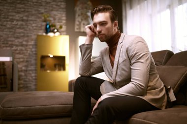 Zarif, şık, gündelik giysiler içinde, orta yaşlı yakışıklı bir adam evinde, kanepede düşünceli bir şekilde oturur. İç mimari.