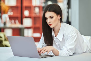 Beautiful young woman using a laptop lying on the bed in her cozy studio apartment. Lifestyle. Home interior, furniture.