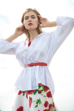 Açık beyaz gömlekli ve çiçekli etekli şık genç bir kadın parlak gökyüzüne karşı duruyor. Yaz tatili.
