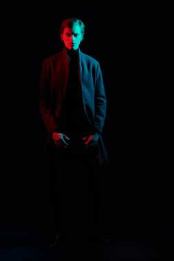 Karanlıkta paltosunun içinde duran yakışıklı bir gencin kırmızı ve yeşil ışıkla aydınlatılmış tam boy portresi. Erkek modası.  