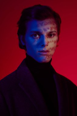 Stüdyoda koyu kırmızı ve mavi ışıkta poz veren zarif koyu renk elbiseli yakışıklı bir adamın portresi. Yüzüne sembollerin gölgeleri düşüyor..