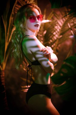 Orman fotoshop 'u. Uzun dalgalı sarı saçları ve parlak makyajlı güzel kız tropik yeşillikler arasında orkide ile bikinili poz veriyor. Spa, cilt bakımı ve kozmetik ürünleri. Tropikal ülkelerde tatil.