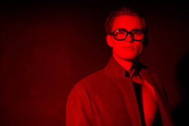 Çekici bir genç adam çıplak bir gövde ve şık gözlüklerle poz veriyor. Koyu kırmızı ışıkta moda portresi. Erkekler için optik.