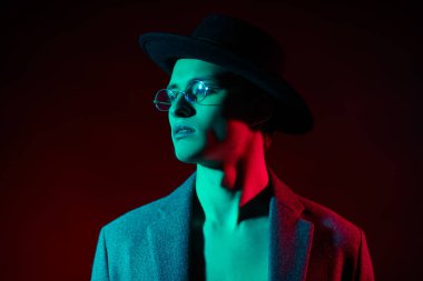 Paltolu, şapkalı ve gözlüklü, koyu kırmızı ve yeşil ışıkta poz veren yakışıklı genç adam. Erkekler için optik. Erkek modası. 