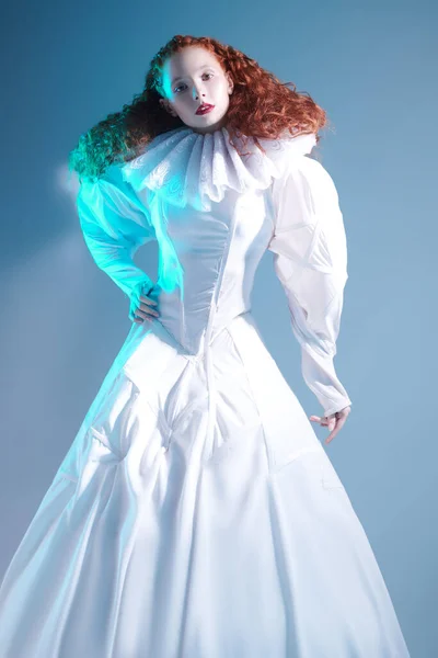 Fashion art history. Sophisticated female model with lush red curly hair posing in a white art dress with late renaissance ruffled collar. Studio portrait on blue bakground in blue .