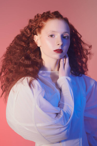 Sophisticated female model with lush red curly hair posing in a white art dress. A studio portrait onpink background in blue light. Fashion art. 
