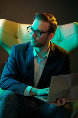 Zarif takım elbiseli ve gözlüklü başarılı iş adamı modern lüks bir iç mekanda deri koltukta oturan bir dizüstü bilgisayarda çalışıyor. Moda. İş dünyası.