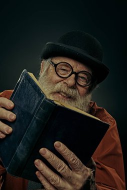 Melon şapkalı ve gözlüklü uzun gri sakallı bilge yaşlı bir adam eski bir kitap okuyor ve gülüyor. Siyah arkaplan. 