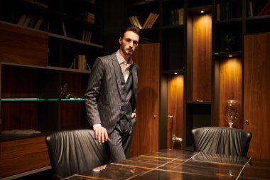 Zarif takım elbiseli yakışıklı esmer adam lüks bir ofiste duruyor. - Mafya. Erkek güzelliği, moda.