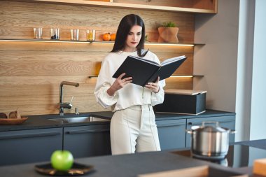 Güzel genç bir kadın yemek kitabını okurken evdeki mutfağında dikiliyor. Sağlıklı beslenme ve beslenme. Modern iç mekan, mutfak mobilyası..