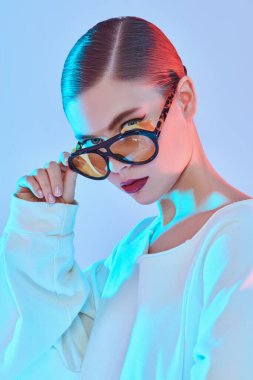 Beyaz uzun kollu gömlek ve mavi ışıkta sarı gözlüklü güzel, şık bir bayan. Kadın aksesuarları, gözlükler..