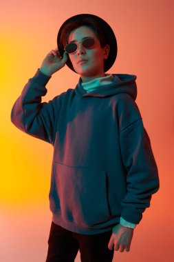 Moda kapüşonlu, şapkalı ve güneş gözlüklü zeki bir çocuk karışık renkli ışık altında poz veriyor. Gençlik modası. Hippi çocuk.. 