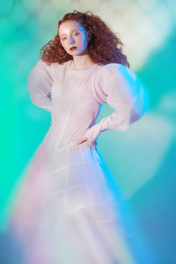 Beyaz bir sanat elbisesi içinde poz veren gür kırmızı kıvırcık saçlı sofistike bir kadın model. Stüdyo portresi. Sis içinde kırmızı bir ışık. Moda sanatı. 