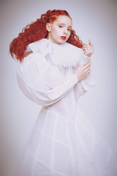 Art portrait of a refined fashion model girl with lush red curly hair posing in a white haute couture dress. Studio shot on light grey background. 