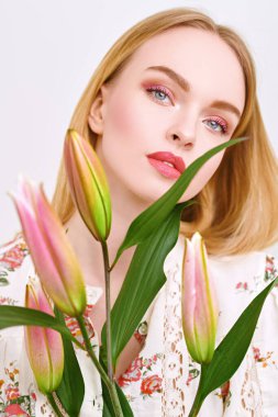 Romantik güzel sarışın kız beyaz arka planda kapalı tomurcuklu zambak çiçekleriyle poz veriyor. Pembe renkli açık renk makyaj. Parfüm ve kozmetik.