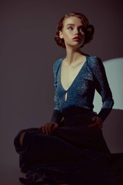 Koyu gümüş elbiseli, saçları ve makyajlı, 20 'li yaşlarda karanlık odada poz veren büyüleyici genç bir kadın. Sanat tasviri. 