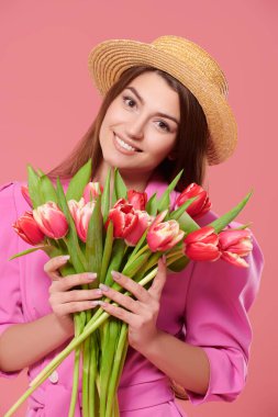 Bahar havası. Pembe elbiseli ve hasır şapkalı güzel kız bir buket lale çiçeği ve gülümseme tutuyor. Anneler Günü. Sevgililer Günü. Pembe arkaplan.