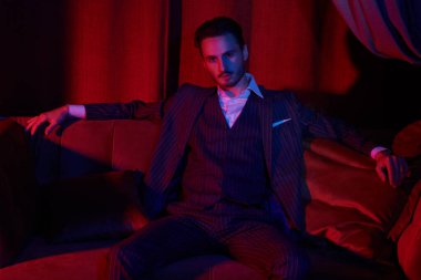 Yakışıklı bir esmer adamın portresi kırmızı ışıkta lüks bir dairede kanepede oturuyor. Göz kamaştırıcı bir yaşam tarzı. Moda fotoğrafı. Erkek kıyafetleri ve aksesuarları..