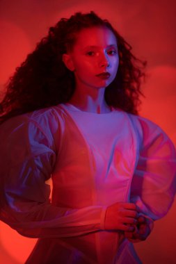 Beyaz bir sanat elbisesi içinde poz veren gür kırmızı kıvırcık saçlı sofistike bir kadın modelin portresi. Karanlık bir odada koyu kırmızı ışıklı bir stüdyo portresi. Moda sanatı. 