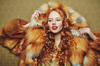 Kızıl saçlı güzel bir kadın açık kahverengi bir arka planda lüks bir tilki kürkünün içinde poz veriyor. Kış güzellik modası. Kürk manto tarzı.
