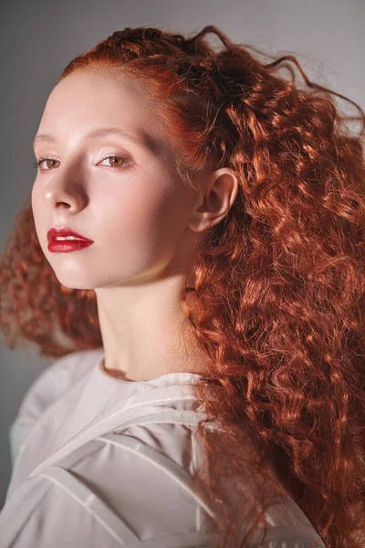 Close up portrait of a refined fashion model girl with lush red curly hair posing in a white haute couture dress. Studio shot on light grey background.