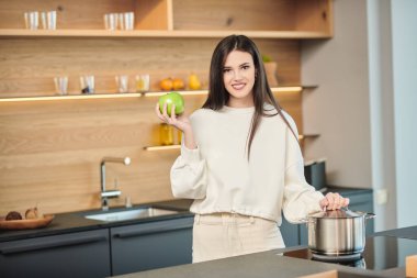 Güzel esmer kız elinde yeşil bir elma tutarak gülümsüyor. Evdeki mutfakta poz veriyor. Sağlıklı beslenme ve beslenme. Modern iç mekan, mutfak mobilyası..