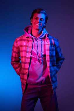 Moda kıyafetleri içindeki genç adam stüdyoda mavi arka planda karışık renkli ışık altında poz veriyor. Gençler için sokak modası.