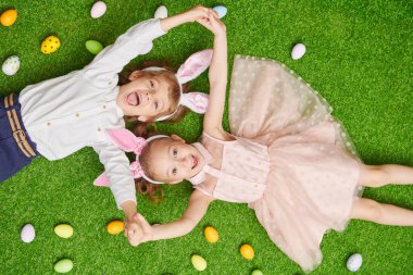 Şirin, gülümseyen bir oğlan ve tavşan kulaklı bir kız renkli yumurtalarla yeşil bir çimenlikte uzanıyorlar. Paskalya tatili.