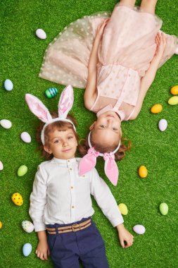 Şirin, gülümseyen bir oğlan ve tavşan kulaklı bir kız renkli yumurtalarla yeşil bir çimenlikte uzanıyorlar. Paskalya tatili.