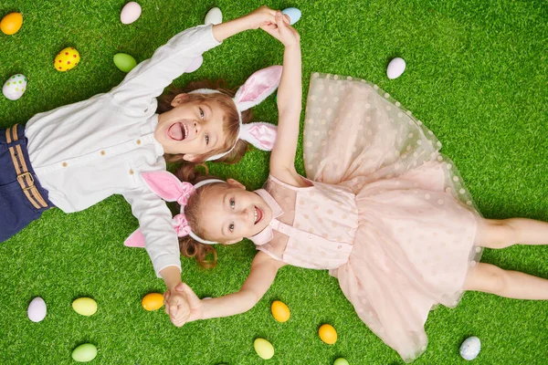 Şirin, gülümseyen bir oğlan ve tavşan kulaklı bir kız renkli yumurtalarla yeşil bir çimenlikte uzanıyorlar. Paskalya tatili.