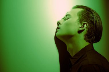 Yakışıklı genç adam, üzgün ve yalnız duruyor, koyu yeşil ışıkta duvara yaslanıyor. Duygular ve duygular. 