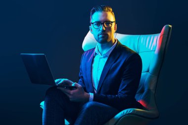 Zarif takım elbiseli ve gözlüklü başarılı iş adamı modern lüks bir iç mekanda deri koltukta oturan bir dizüstü bilgisayarda çalışıyor. Mavi ışıkta moda. İş dünyası.