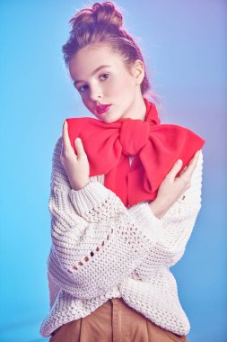 Beyaz kazaklı zarif bir kızın portresi ve boynunda büyük kırmızı bir fiyonk yumuşak pembe ve mavi bir ışığa karşı. Güzellik, moda konsepti. Kış tarzı.