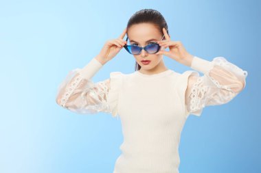 Zarif beyaz bluz ve açık mavi arka planda güneş gözlüğü takan güzel bir yaz kızı. Yaz modası ve makyaj. Optikler. 