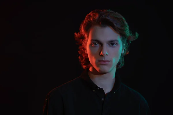 Portrait of a handsome young man with wavy blond hair on a dark background with mixed color light and shadows. Men's beauty.