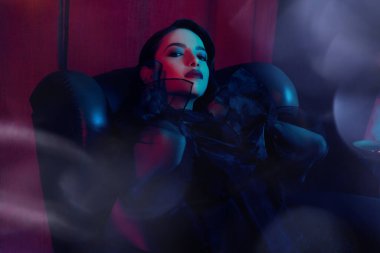 Moda şıklığı. Şık siyah elbiseli güzel esmer kız lüks bir dairede deri koltukta oturuyor. Güzellik, moda konsepti. Göz kamaştırıcı yaşam tarzı.