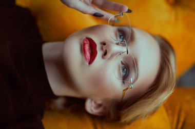 Sarı bir kanepede uzanan zarif siyah elbiseli ve gözlüklü güzel moda hanımefendisi. Güzellik, moda. Optik biçimi.