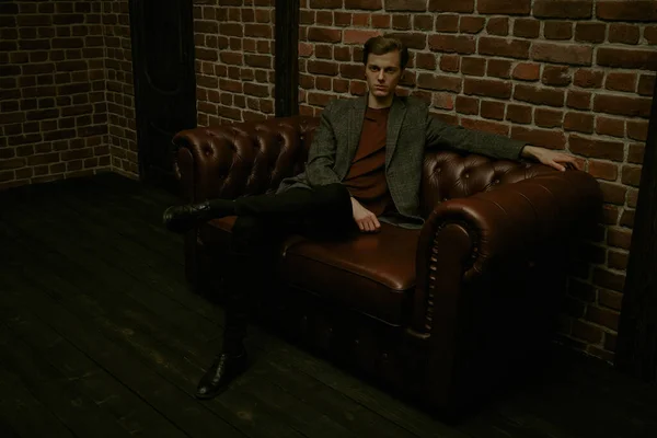 A handsome guy sits on a leather sofa in a dark loft-style room. Men\'s beauty, fashion. Lifestyle.