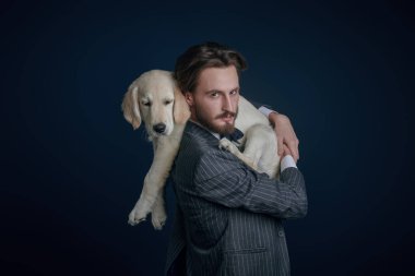 Erkek modası. Zarif takım elbiseli yakışıklı bir adamın portresi. Siyah arka planda Golden Retriever köpeğini tutuyor.. 