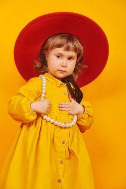 Şirin küçük kız modası. Sarı arka planda, kırmızı şapkalı ve sarı elbiseli sevimli küçük bir kızın portresi..