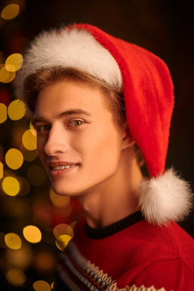 Портрет счастливого красивого молодого человека в шляпе Санта Клауса, празднующего Рождество. Фон сверкающих праздничных огней. Копирование пространства.