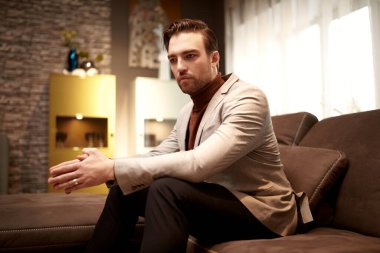 Zarif, şık, gündelik giysiler içinde, orta yaşlı yakışıklı bir adam evinde, kanepede düşünceli bir şekilde oturur. İç mimari.