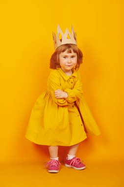 Küçük prenses kız. Kartondan bir taç takmış, kameraya bakıp gülümseyen sevimli bir kızın uzun bir portresi. Mutlu bir çocukluk. Çocukların duyguları ve hayal gücü. Sarı arkaplan. 