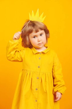 Prenses tacıyla oynayan ve gülümseyen tatlı küçük bir kız. Mutlu bir çocukluk. Çocukların duyguları ve hayal gücü. Sarı arka planda stüdyo resmi. 