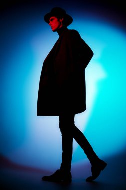 Zarif şapkalı ve ceketli yakışıklı genç adam stüdyoda karışık renkli ışıkta hareket halinde poz veriyor. Sanat portresi. Erkek güzelliği, moda.