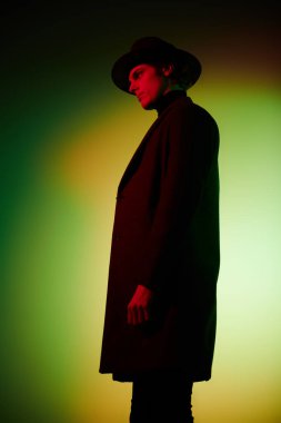 Zarif şapkalı ve ceketli yakışıklı genç adam stüdyoda karışık renkli ışıkta poz veriyor. Sanat portresi. Erkek güzelliği, moda.