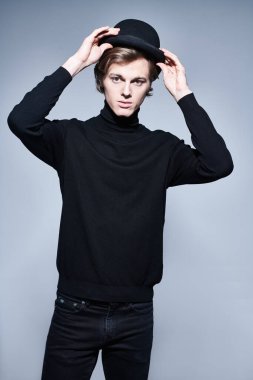 Siyah balıkçı yaka ve gri arka planda melon şapkalı zarif bir gencin portresi. Erkek güzelliği. Moda.. 