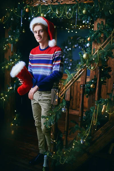 Happy handsome young man in a sweater and Santa Claus hat stands with a x-mas sock at home. Beautiful x-mas decorations. Christmas and New Year.