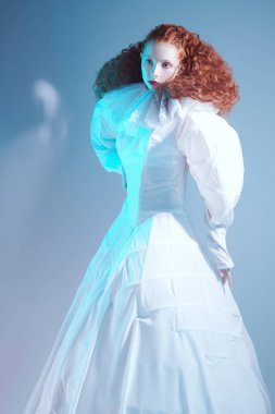 Mavi arka planda beyaz bir sanat elbisesi içinde poz veren gür kırmızı kıvırcık saçlı güzel bir kadın model. Stüdyo portresi. Sis içinde kırmızı bir ışık. Moda sanatı. 