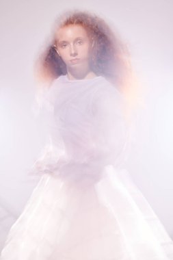 Saf kırmızı kıvırcık saçlı, parıltılar ve sis arasında uzun beyaz bir elbise giymiş zarif bir kadın modelin portresi. Sanat modası. 